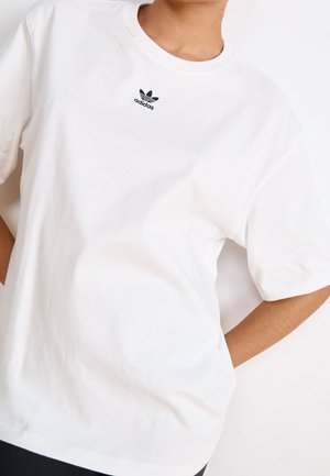 Osoba nosząca prostą białą koszulkę marki Adidas z małym czarnym logo Adidas umieszczonym centralnie, blisko kołnierza.