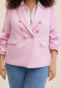Femme portant un blazer croisé rose clair à boutons dorés et une chemise blanche, associée à un jean bleu, debout les bras détendus.