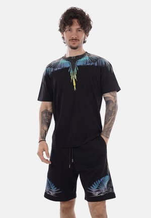 Hombre con cabello rizado que lleva una camiseta negra y pantalones cortos con diseños de alas de aves azules y amarillas, de pie con una mano en el bolsillo.