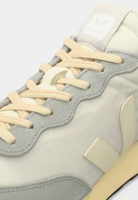 Αθλητικό παπούτσι ανοιχτού γκρι χρώματος με υφή από καστόρι, μπεζ λεπτομέρειες και κρέμ κορδόνια. Διαθέτει καουτσούκ σόλα και έντονο λογότυπο στη γλώσσα του παπουτσιού.