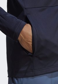 Granatowa koszula z długim rękawem z boczną kieszenią, o gładkiej fakturze i minimalistycznym wzornictwie, z widocznymi detalami szwów i otwarciem.