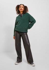 Maglione verde con texture e scollo a V, indossato su una maglietta nera, abbinato a pantaloni di pelle neri e sneakers nere. Aspetto casual.