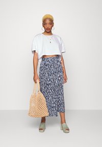 Camisa branca curta, saia maxi azul com padrão, saltos verde-escuro e uma mala de vime bege. As joias incluem brincos em argola e colares em camadas.