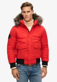 Chaqueta acolchada roja con capucha forrada de pelo, que presenta dos bolsillos frontales y puños acanalados. Detalles con cremallera negra y tejido texturizado.