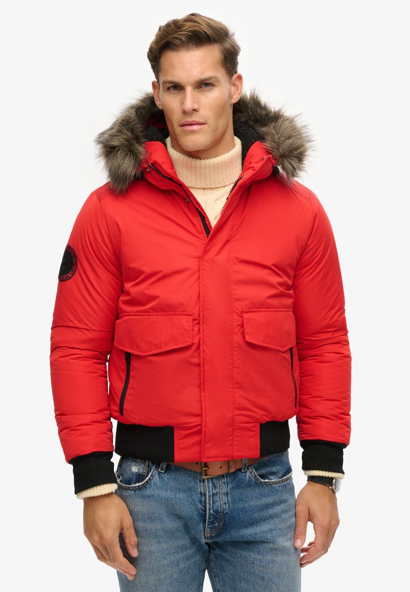 Veste rembourrée rouge avec une capuche en fourrure, dotée de deux poches avant et de poignets côtelés. Accents de fermeture éclair noire et détails en tissu texturé.