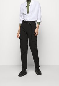 Zwarte cargobroek met hoge taille, verstelbare riem en meerdere voorkruiden. Gecombineerd met een wit overhemd en een groene ribbel top.
