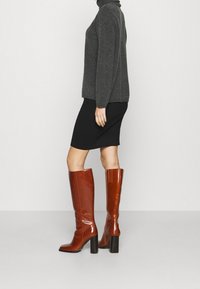 Bruna läder knälånga stövlar med fyrkantig tå, blockklack och vertikal dekorationsdetalj. Bärs med en grå polotröja och svart kjol.