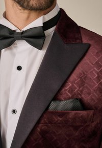 Sacou de smoking din catifea burgundă, cu rever din satin negru, cămașă albă și batista de buzunar cu model. Papion negru și buton vizibile.