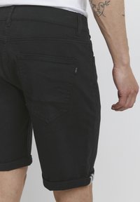 Crni traper kratke hlače s preklopljenim rubovima, s leđnim džepom s detaljima šivanja i malom oznakom branda sa strane. Teksturirana tkanina.