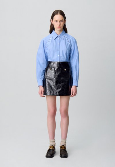 Camisa a rayas azul claro con detalles de perlas, combinada con una falda de cuero negro brillante que cuenta con dos bolsillos y botones dorados. Zapatos y calcetines marrones.