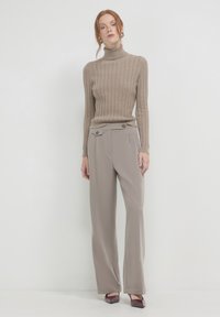 Beige gerippter Rollkragenpullover kombiniert mit hellgrauen weiten Hosen, die zwei vordere Knöpfe und eine glatte Textur aufweisen. Dunkle Absätze runden den Look ab.