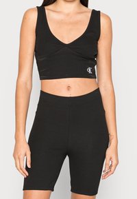Kvinna som bär en svart ribbad crop top och högmidjade cykelbyxor med en liten vit CK-logotyp upptill.