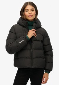 Svart, vadderad pufferjacka med huva, zippered framsidan, elastiska manschetter och ribbstickad detalj vid kragen. Logotyp på ärmen.
