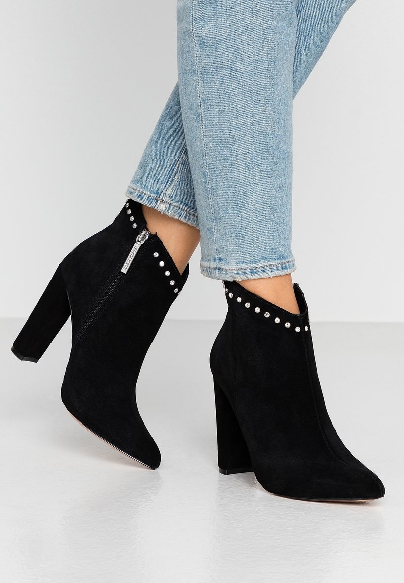 Persona che indossa jeans azzurri chiari e stivali neri in suede con tacco alto, alla caviglia, dotati di zip laterale e dettagli in borchie argentate lungo il bordo superiore.