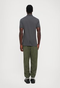 Man standing facing away wearing a dark gray short-sleeve polo shirt, olive green patterned pants, white socks, and black shoes.