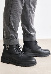 Svarte snørestøvler i kampsjorte stil med glatt overflate, tykk såle og detaljer i metall, kombinert med gråstripete bukser.