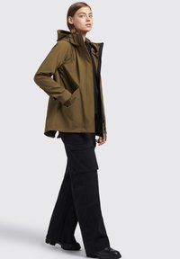 Giacca impermeabile verde oliva con cappuccio, chiusura con zip e bottoni a pressione, due tasche laterali, abbinata a pantaloni cargo neri a gamba larga e stivali.