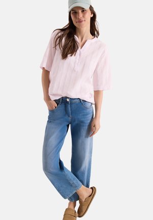 Femme portant un chemisier rose clair à manches courtes, un jean large bleu clair, des chaussures marron à enfiler et une casquette de baseball blanche, debout avec une main dans la poche.