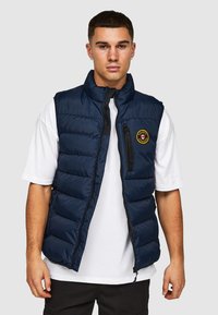 Gevoerde marineblauwe vest met hoge kraag, ritssluiting aan de voorkant en logopatch op de borst; gecombineerd met een wit T-shirt. Glad textuur en casual ontwerp.