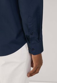Camisa de manga larga azul marino con puños abotonados, que presenta una textura suave y un dobladillo redondeado, combinada con pantalones blancos.