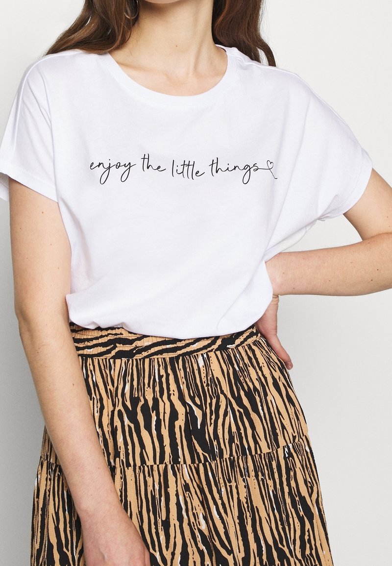 Femme portant un t-shirt blanc avec le texte « profitez des petites choses » et une jupe beige à motifs noirs, main posée sur la hanche.