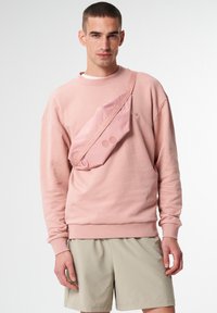 Rosa Sweatshirt aus weichem Stoff mit lässiger Passform, kombiniert mit beigen Shorts. Eine rosa Umhängetasche wird quer über die Brust getragen und verfügt über kreisförmige Akzente.