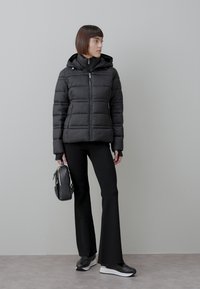 Femme portant une doudoune noire, un pantalon noir évasé, des baskets noires à semelles blanches, tenant un sac à main noir, debout devant un mur gris.