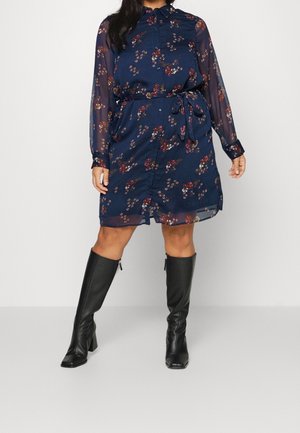 Robe florale bleu marine avec manches longues en tissu transparent, fermeture boutonnée à l'avant, ceinture à la taille et coupe droite. Associée à des bottes noires en cuir montant jusqu'aux genoux.