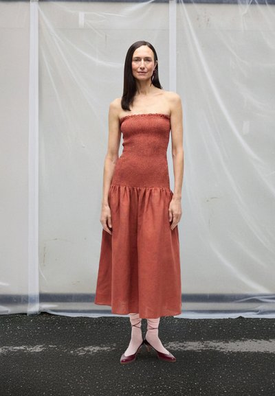 Stroppeløs midikjole i rustfarget, rynket stoff som flommer ut i et samlet skjørt. Brukes med rosa knelange sokker og burgunderrøde hæler.