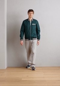 Cazadora bomber verde con rayas blancas en los puños y el dobladillo, pantalones grises y zapatillas negras. Fondo liso con suelo de madera.