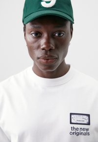 Hvit t-skjorte med et grafisk motiv av en måler og teksten "de nye originalene," kombinert med en grønn caps med et hvitt logo.