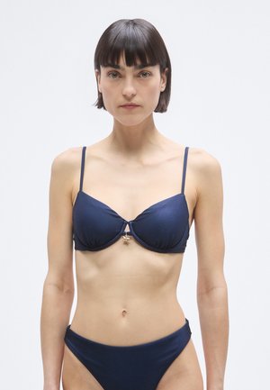 Námořnicky modrý horní díl bikin s nastavitelnými tenkými ramínky, podprsenkovou kosticí a malým zlatým hvězdným detailem uprostřed. Hladká, lesklá textilní struktura.