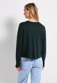 Ciemnozielona bluzka z długim rękawem i okrągłym dekoltem; ma krótki fason i miękką teksturę, zestawiona z jasnoniebieskimi dżinsami z wysokim stanem.