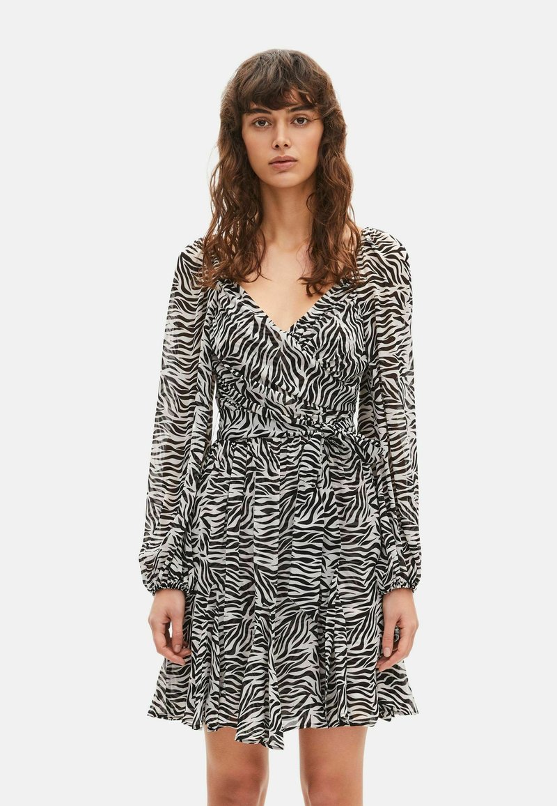 Mujer vestida con un vestido de manga larga con estampado de cebra en blanco y negro, escote en V y cintura fruncida, de pie contra un fondo liso.