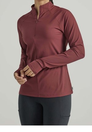 Personne portant un pull-over bordeaux à manches longues côtelées avec un col court à fermeture éclair et des ouvertures pour les pouces, associé à un pantalon noir ajusté.