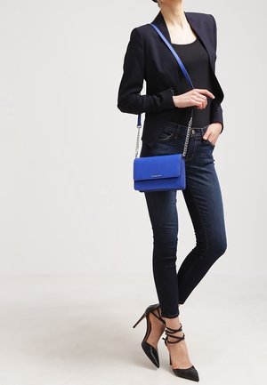 Sininen olkalaukku ketjuhihnalla, suorakulmainen muoto, sileä pinta, yhdistetty tiukkaan mustaan paita ja tummiin skinny-farkkuihin.