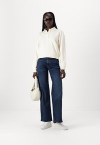 Felpa con colletto crema, jeans dritti blu scuro, sneakers bianche e una borsa bianca. Modella con occhiali da sole neri, texture piatta.