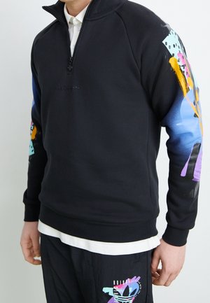 Mann trägt schwarzen Adidas-Pullover mit bunten abstrakten Designs auf den Ärmeln und passenden schwarzen Hosen mit ähnlichen Mustern.