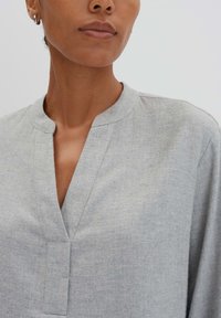 Blouse gris clair avec un col à encolure en V, texture douce et manches larges ; présente des détails de couture minimaux.