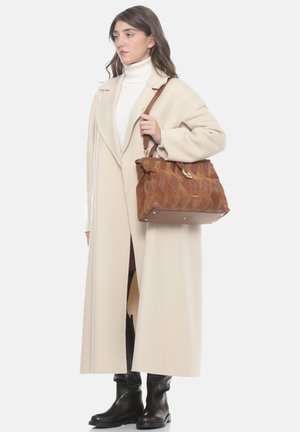 Donna che indossa un lungo cappotto beige e stivali neri, con una grande borsa in pelle marrone con motivo geometrico appoggiata sulla spalla.