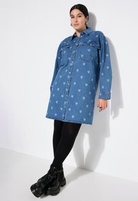 Denimhemdkleid in Blau mit hellblauen Herzmustern, langen Ärmeln, Knopfleiste und zwei Brusttaschen. Getragene schwarze Plateauschuhe.