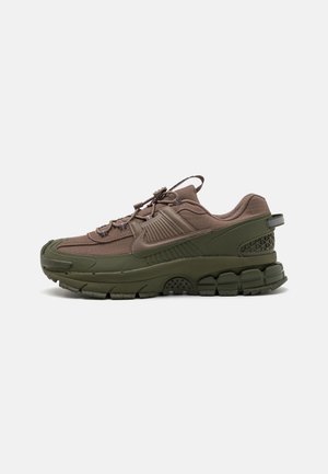 Chaussure de sport marron et vert olive avec semelle texturée, cordon de serrage, languette de traction au talon et logo en forme de swoosh sur le côté, sur fond blanc.