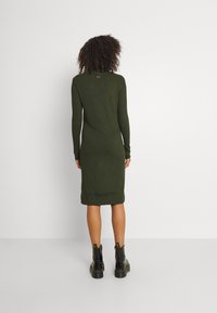 Abito a maglia verde oliva con maniche lunghe e siluetta aderente, caratterizzato da un logo sul colletto e un orlo a metà polpaccio. Abbinato a stivali neri.