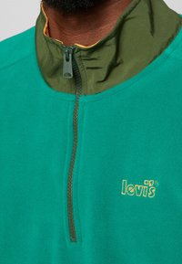Sudadera de forro polar verde con un diseño de medio zipper, cuello verde oliva en contraste y forro interior amarillo. Presenta el logo de "Levi's" en dorado.