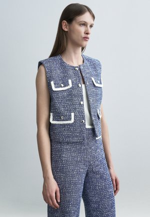 Žena v modrej bezrukávovej vestě z tkaniny s bielym lemovaním a gombíkmi, v kombinácii s pásovými vysokými nohavicami z rovnakého materiálu, stojí pred sivým pozadím.