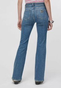 Jean évasé bleu foncé à taille haute, en denim. Il comporte cinq poches et une étiquette logo en métal. Ourlet effiloché en bas.