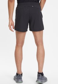 Pantalones cortos deportivos negros con una textura suave, cintura elástica y un bolsillo trasero discreto. Llevados con zapatillas de correr grises con patrón.