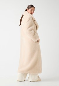 Donna con lunghi capelli scuri indossa un lungo cappotto teddy color crema, pantaloni di maglia color crema e pantofole, mentre è in piedi contro uno sfondo bianco.