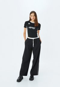 Sort cropped t-shirt med hvide detaljer og "SPEED" logo, parret med løse sorte bukser med snøre og hvide sidelister.