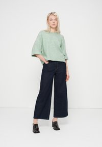 Haut oversize vert clair texturé avec manches larges, associé à un pantalon large en denim foncé et des chaussures à talons noires brillantes.